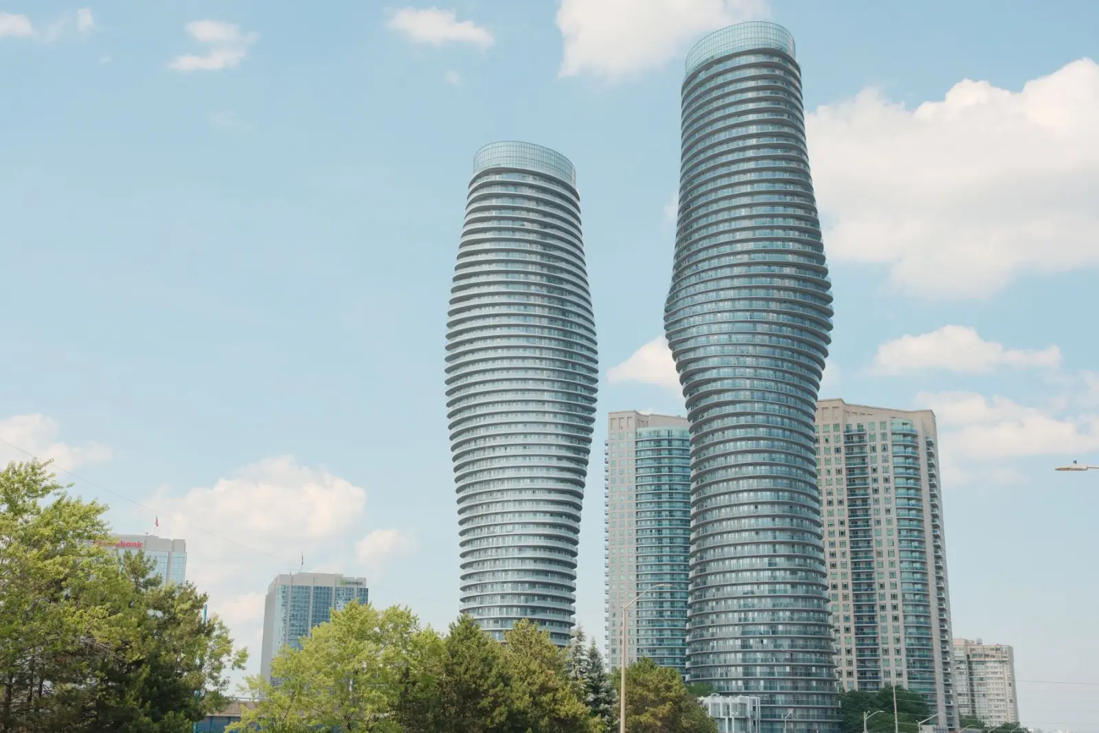 Mississauga's curving Absolute World towers rise above trees against a blue, cloud-dotted sky.