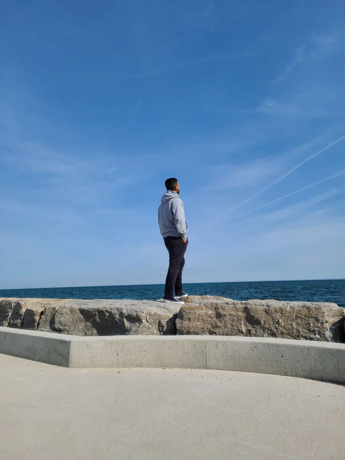 A man stands on the stone breakwater of the Oakville lakeshore, looking out over blue Lake Ontario.