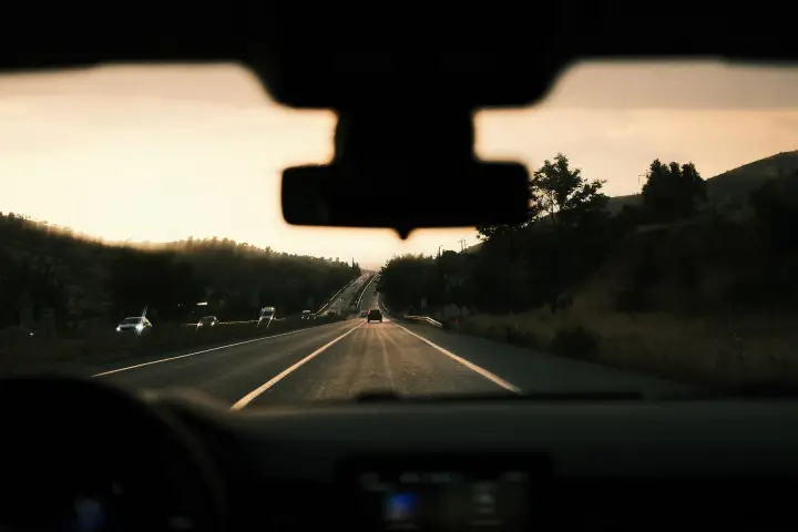 A highway at sunset seen from the driver's seat, through the windshield, with the rear-view mirror mount at the top of the glass.