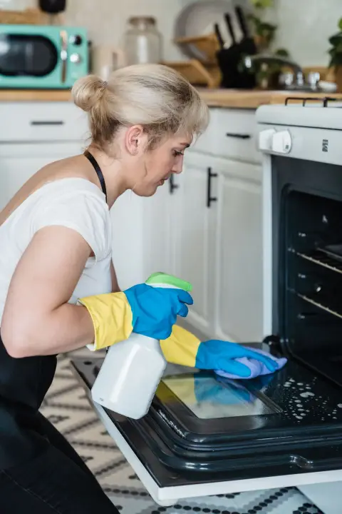 A woman in rubber gloves sprays and wipes the inside of an open oven door in a white kitchen.