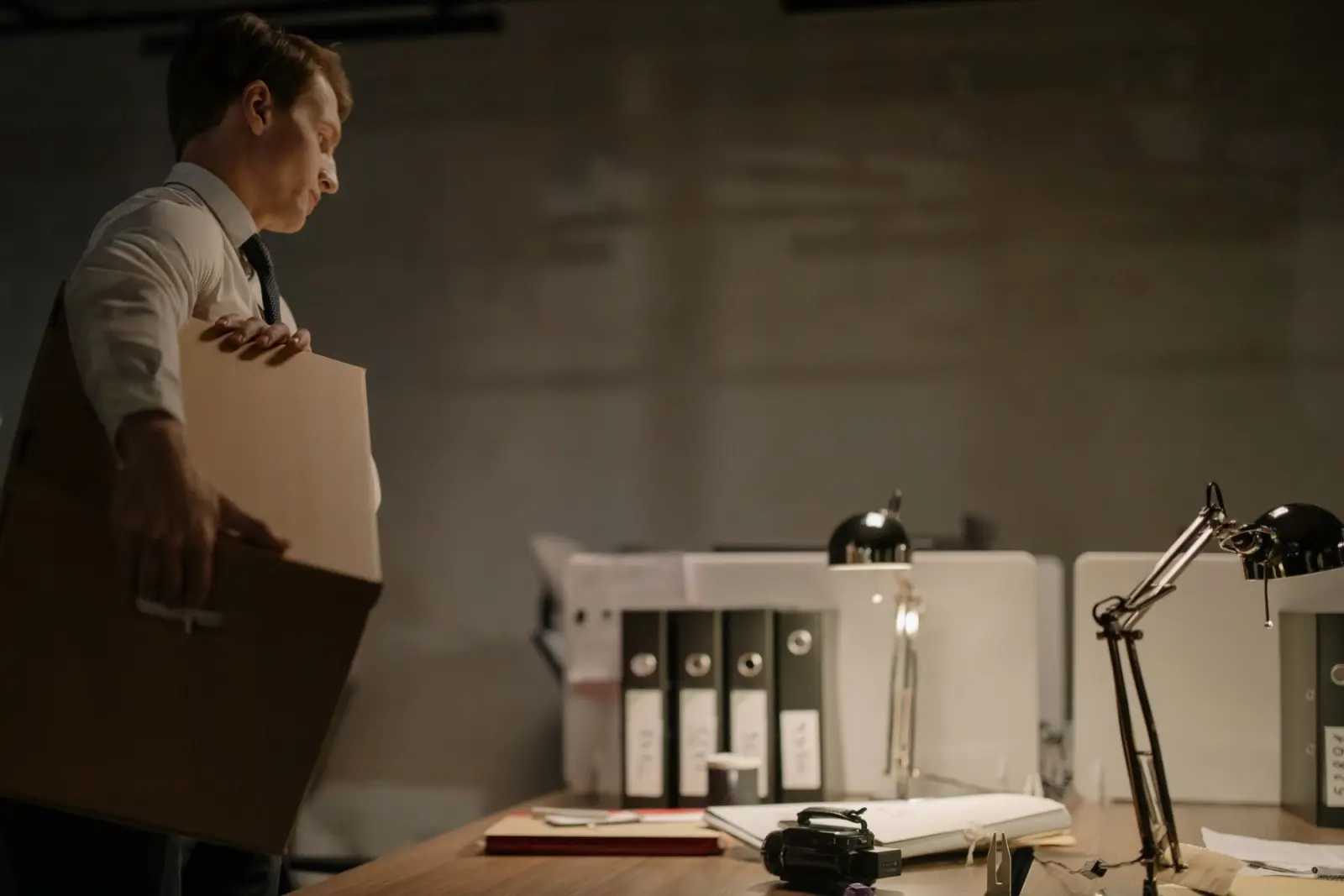 A man in a shirt and tie carries a cardboard box past an office desk with binders and desk lamps.